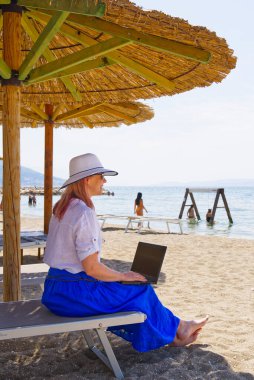 Herhangi bir yerden çalış. Genç gülümseyen kadın, hasır şapkalı kadın plajda dizüstü bilgisayarla çalışıyor..