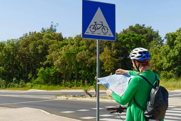 Τουριστικό κορίτσι σε ένα κράνος σε ένα ποδήλατο εξετάζει ένα χάρτη στη διασταύρωση των δρόμων ποδήλατο — Φωτογραφία Αρχείου