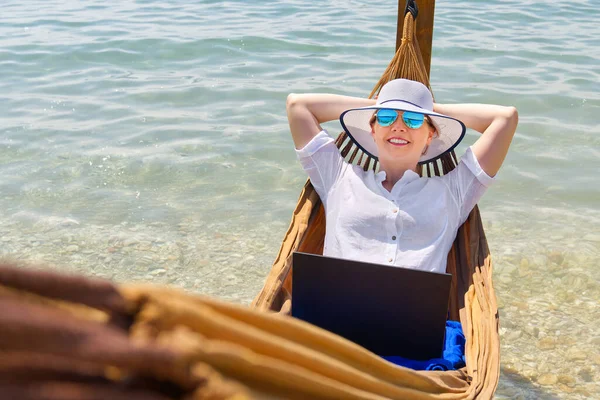 Práce odkudkoliv. Mladý smajlík žena, žena na volné noze v slamáku pracuje na notebooku na pláži. — Stock fotografie