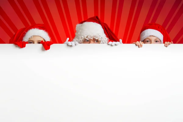 Satna claus con niños divertidos en sombreros de gnomo de Navidad rojos asomándose detrás de una pancarta blanca en blanco — Foto de Stock