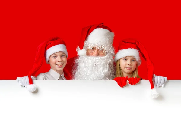 Satna claus mit lustigen Kindern in roten Weihnachtszwergenhüten, die hinter einem weißen Transparent hervorlugen — Stockfoto