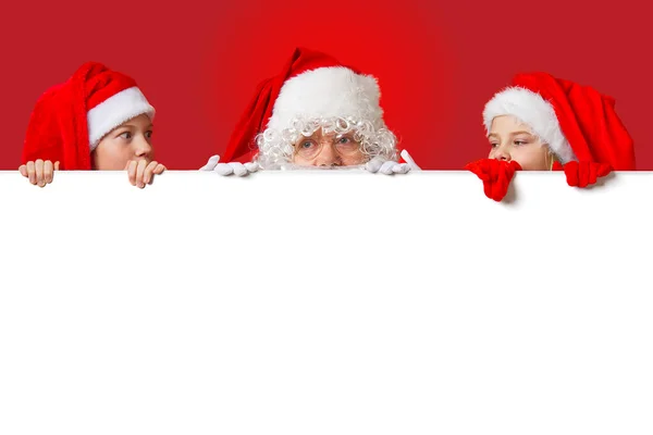 Satna claus con niños divertidos en sombreros de gnomo de Navidad rojos asomándose detrás de una pancarta blanca en blanco — Foto de Stock