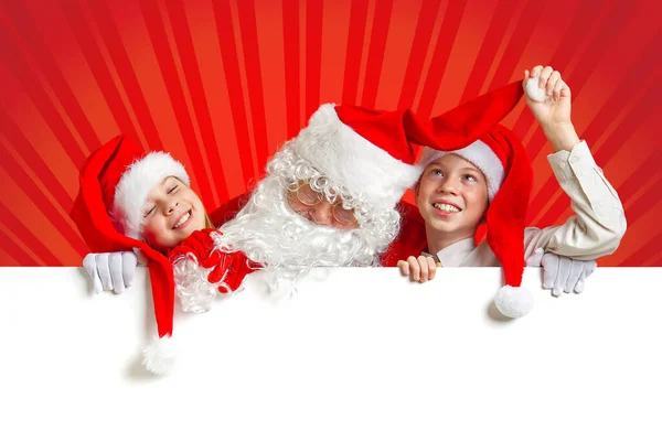 Satna claus mit lustigen Kindern in roten Weihnachtszwergenhüten, die hinter einem weißen Transparent hervorlugen — Stockfoto