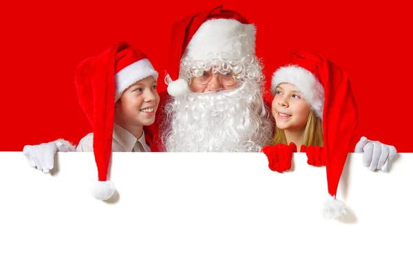 Satna claus avec des enfants drôles en chapeaux de gnome de Noël rouge jetant un coup d'oeil derrière une bannière blanche vierge — Photo