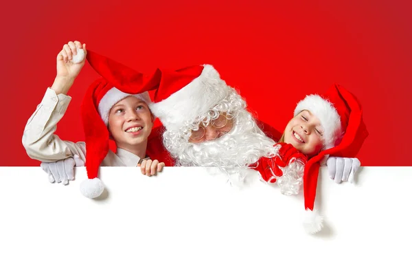 Satna claus mit lustigen Kindern in roten Weihnachtszwergenhüten, die hinter einem weißen Transparent hervorlugen — Stockfoto