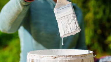 Bir insan boya fırçasını uygun bir kovadan beyaz boyaya batırır. Bu resim ev geliştirmesini ve DIY projelerini sembolize ediyor, yenileme ve sanatsal uygulamaları göstermek için mükemmel.