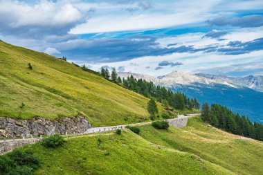 Alpler 'deki dolambaçlı bir dağ yolunun çarpıcı güzelliğini deneyimleyin, yemyeşil tepeler ve dramatik bir gökyüzünün altında yükselen zirveler, seyahat ve turizm için ideal..