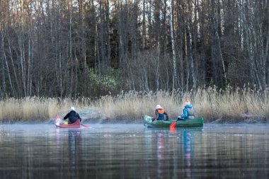 Sazlıklar ve ormanla çevrili sakin bir gölde kano yapan üç kişi. Huzurlu sular sonbahar manzarasını yansıtır, barışçıl bir açık hava macera sahnesi yaratır..