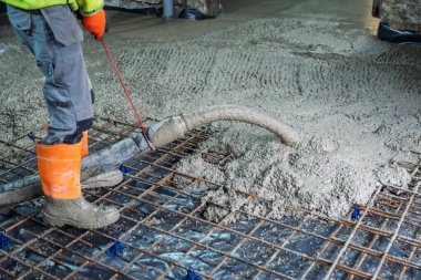 Turuncu çizmeli ve eldivenli bir inşaat işçisi inşaat alanındaki demirin üzerine beton döküyor. İşçi düz bir yüzey oluşturmak için betonu düzleştiriyor..