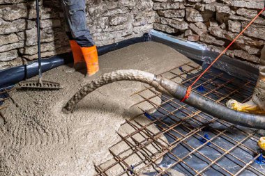 Bir inşaat işçisi, el arabası ve aletlerle yeni bir bodrum katı döşemesi için beton döküyor. Sahne, sağlam ve pürüzsüz bir yüzey için demiri destekler ve detaylara dikkat eder..
