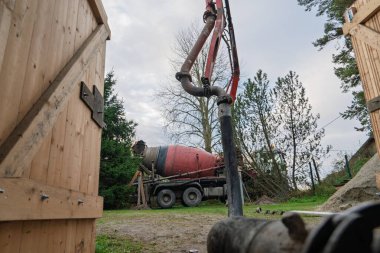 Bir çimento kamyonu inşaat alanındaki bir patlamaya beton pompalıyor. Alanı çevreleyen tahta yapılar, devam eden bir inşaat projesini gösteriyor, endüstriyel faaliyetleri gösteriyor..