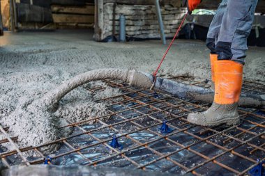 İnşaat işçisi bir bina temeli için beton döküyor. Yapısal bütünlük için güçlendirme çelik şebekesi yerleştirildi. Güvenlik teçhizatı bölgede güvenliğin önemini vurguluyor.