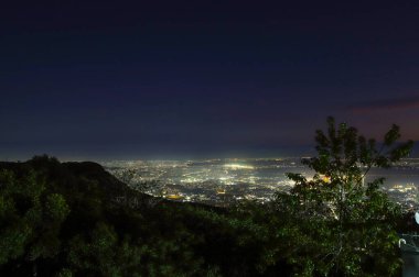 Geniş açılı bir gece çekimi yüksek bir dağ zirvesinden geniş bir şehir manzarasını yakalıyor. Aşağıdaki şehir, ön plandaki karanlık ağaç siluetleriyle ve yukarıdaki derin mavi gökyüzüyle çelişen parıldayan yapay ışıklarla dolu bir deniz.