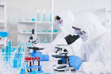 Antiveba kıyafeti giyen bir tıp uzmanı virüsten tamamen korunur ve steril lastik eldiven giyer ve laboratuarın içinde otururken mikroskop merceğine bakar..