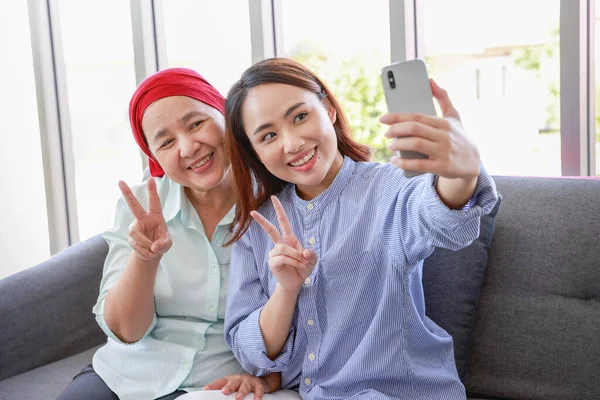 A senior woman with cancer wearing a headscarf relaxes at home with her adult daughter and take picture by smartphone in the living room.The women are full of hope for the future.
