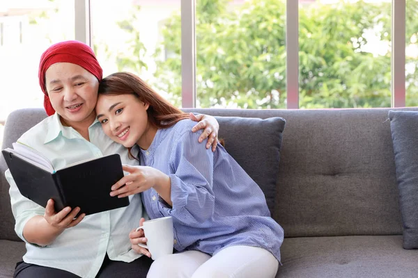 A senior woman with cancer wearing a headscarf relaxes at home with her adult daughter and reading a book with smiling.The women are full of hope for the future