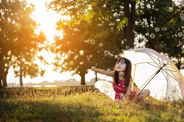 日落时带着漂亮的彩色雨伞 坐在公园绿草上的可爱的小女孩 — 图库照片