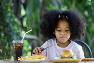 childhood and eating concept - little african american curly hair girl holding knife and looking at burger and french fries on the table .enjoying unhealthy food.