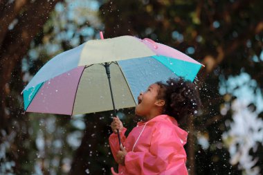 Mutlu komik çocuk, sonbahar duşunun altında şemsiyeli siyah saçlı kıvırcık saçlı. Kız pembe yağmurluk giyiyor ve yağmurun tadını çıkarıyor. Doğada oynayan çocuk..