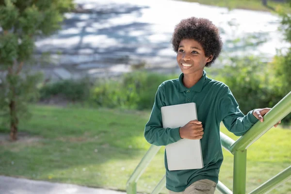 一个黑发卷曲的非洲男孩抱着笔记本电脑 微笑着看着学校大楼栏杆下的摄像机 看着摄像机 — 图库照片