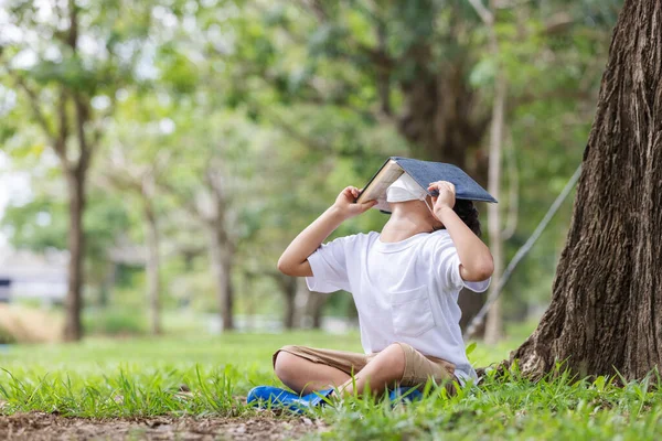 一个戴着卫生面具的白人男孩一个人坐在一棵树下看书盖头 — 图库照片