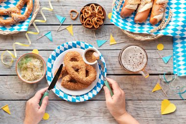 Oktoberfest geleneksel yiyecek ve bira, düz masa üzerinde mavi ve beyaz kağıt Bavyera süslemeleri ile. Bira bardağıyla el ele tutuşmak. Geleneksel festival hamur işleri: simit ve ekmek çubukları.