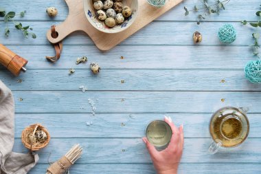 Mavi ahşap masada sıfır atık Paskalya arkaplanı. Bıldırcın yumurtası, doğal bahar süslemeleri, dallar ve okaliptüs. Çaydanlıkta yeşil çay. Çayla el ele tutuşun..