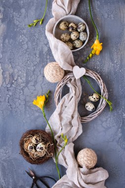 Paskalya düzlüğü, kuş yuvasında bıldırcın yumurtaları, keten tekstil ve çevresi ile koyu bir zemin üzerinde uzanıyordu. Koyu renk masa üzerinde sarı frezya çiçekleri, makas ve rattan çelengi. Plastiksiz Paskalya süslemeleri.