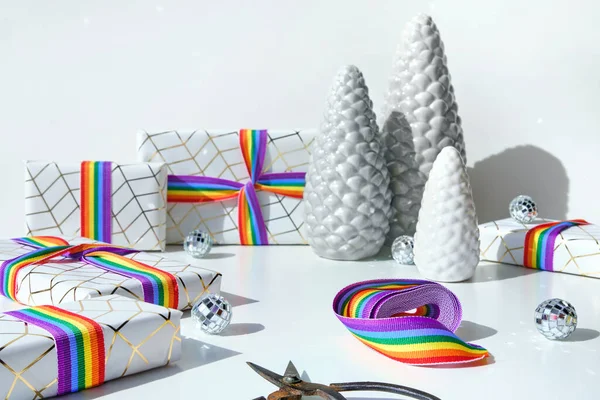 Christmas gifts with rainbow ribbon in LGBTQ community flag colors. Xmas decorations - ceramic fir trees, red baubles, mirror disco balls, LGBT rainbow ribbon and scissors.