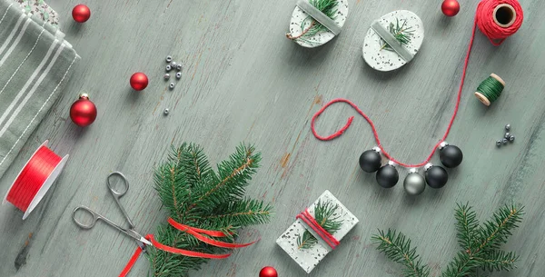 Panoramic Christmas background on grey wood in white and red. Geometric flat lay with toys, flowers, gift boxes with natural handmade decorations with ribbon and winter green leaves.