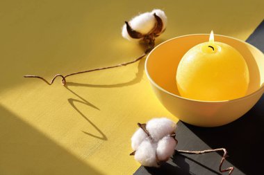 Aydınlatıcı Sarı ve Ultimate Gri, 2021 yılının renkleri. Dekoratif tepside yanan mum ve bölünmüş sarı gri arka planda pamuk çiçeği. Güneş ışığı ve uzun gölgeler.