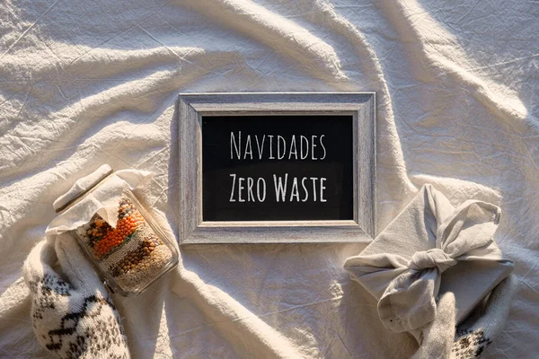 Blackboard with text Navidades Zero Waste in Spanish, that means Zero Waste Christmas. Hands in gloves with eco friendly gifts for winter holidays. Flat lay on light linen textile background.