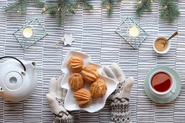Creative Xmas table setup for tea with cakes. Creative flat lay with yummy eclairs or brewing cakes. Christmas tree shape plate with cakes. Stripy textile tablecloth. Fir twigs with light garland.