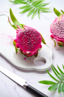 Pembe ejderha meyvesi, pitaya veya pitahaya beyaz tabakta küp şeklinde kesilmiş. Moda süper yiyecek malzemesi. Kesme tahtası ve bıçakla yakın plan görüntü. Egzotik palmiye yaprakları.