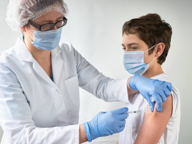 Coronavirus, grip ya da kızamık aşısı konsepti. Sıhhiyeci, doktor, hemşire, sağlık uygulayıcısı genç çocuğu şırıngada aşı ile aşıladı. Üniforma, kulübe ve eldiven giyiyor. İkisi de yüz maskesi takıyor.