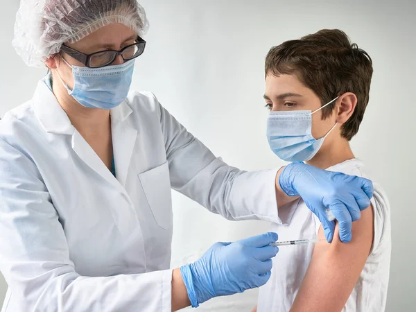 Coronavirus, flu or measles vaccine concept. Medic, doctor, nurse, health practitioner vaccinates teenage boy with vaccine in syringe. She is wearing uniform, hut, gloves. Both people wear face mask