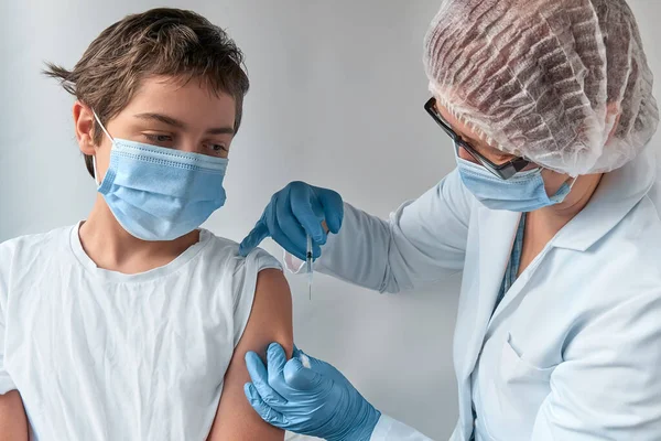 Coronavirus, flu or measles vaccine concept. Medic, doctor, nurse, health practitioner in white gown and face mask vaccinates teenage boy with vaccine in syringe.