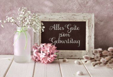 Springtime background. Spring flowers and a blackboard on white table. Pink hyacinth, baby breath and pussy willow. Text Alles gute zum Geburtstag means Happy Birthday. clipart