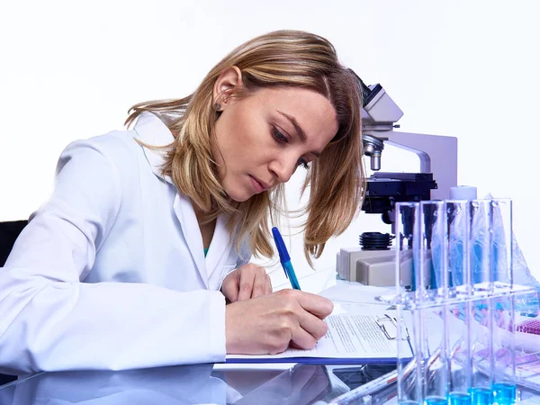 Young female scientist or tech writes report or lab journal in modern laboratory with microscope, glass tubes with samples and timer, This image is toned.