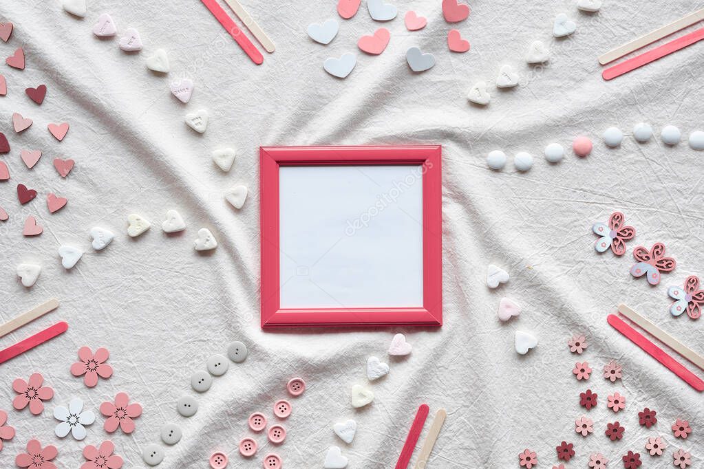 Abstract Springtime background. Pink picture frame with copy-space. Flat lay with hearts, candy, buttons, small decor arranged on textile background. Off white tablecloth.