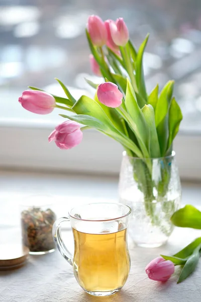 グラスマグカップでお茶と春の背景 リネンのテーブルクロスとテーブルの上にピンクのチューリップの束 窓からの日差し ガラス貯蔵瓶の中の乾燥茶葉 — ストック写真