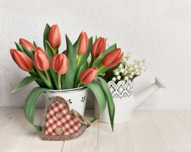 Paskalya ya da bahar doğum günü kutlaması. Basit bir tebrik kartı tasarımı. Su kutusunda bir sürü lale. Ahşap ve tekstilden yapılmış dekoratif bir kalp. Beyaz arka plan doğal, duvarın yanındaki tahta masa..