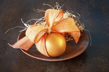 Turuncu yaylı tek bir altın Paskalya yumurtası. Bahar dekorasyonları, kahverengi arka planda koyu kırsal tabakada saman. Basit Paskalya tebrik kartı tasarımı.