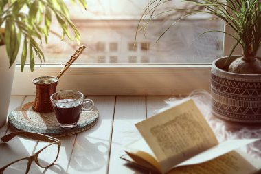 Geleneksel Türk bakır kahve makinesinde pişirilmiş doğu kahvesi. Pencere kenarında çiçekler. Egzotik bitkiler ve klasik kitapla ahşap pencere pervazı. Sade sahne, hygge konsepti. Soğuk sonbahar, kış günü.
