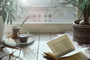 Geleneksel Türk bakır kahve makinesinde pişirilmiş doğu kahvesi. Pencere kenarında çiçekler. Egzotik bitkiler ve antika kitapla ahşap pencere pervazı. Sade sahne, hygge konsepti..