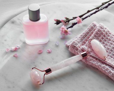 Kristal gül kuvars yüz masajı için. Pembe cilt bakım ürünü ya da buzlu cam kavanozdaki parfüm. Beyaz mermer ve tekstil arka planı, doğal ışık. Bahar kiraz çiçekleri.