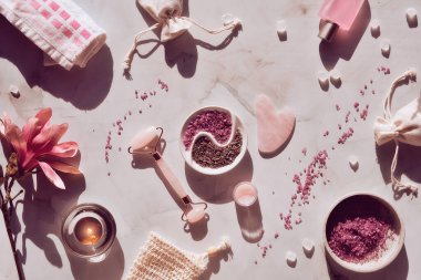 Kristal gül kuvars yüz silindiri ve gua sha taşı güzellik yüz masajı terapisi için, düz mermer masa üzerinde manolya çiçekleri, tonlu görüntü. Uzun gölgeler ve mum. Gerekli yağ şişesi.
