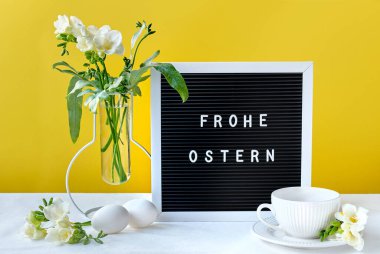 Frohe Ostern 'a Mutlu Paskalyalar demek. Bahar Frezya çiçekleri ve çay fincanı olan bir mektup tahtası. Üzerinde yazı olan siyah bir yazı tahtası. Doğal Paskalya yumurtaları, beyaz çay fincanları ve aydınlatıcı sarı arka planda çiçekler..