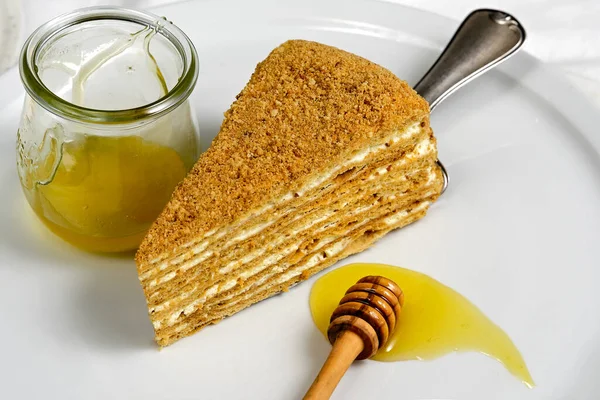 Russian honey cake slice on white plate on off white textile tablecloth. Traditional 8 layer delicious dessert from former Soviet Union. Honey in small jar with wooden stick.