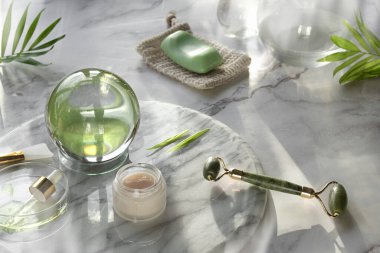 Yeşil yeşim yüzlü silindir, masaj aleti ve mermer taşlı arka planda yüz bakım ürünleri. Cam kristal küre ve egzotik palmiye yaprakları. Havlu, cilt toniği, temel yağ, serum..